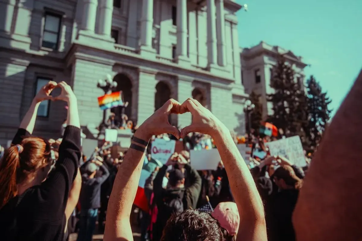 A crowd at a rally outside a state capitol, hands raised in heart shapes.