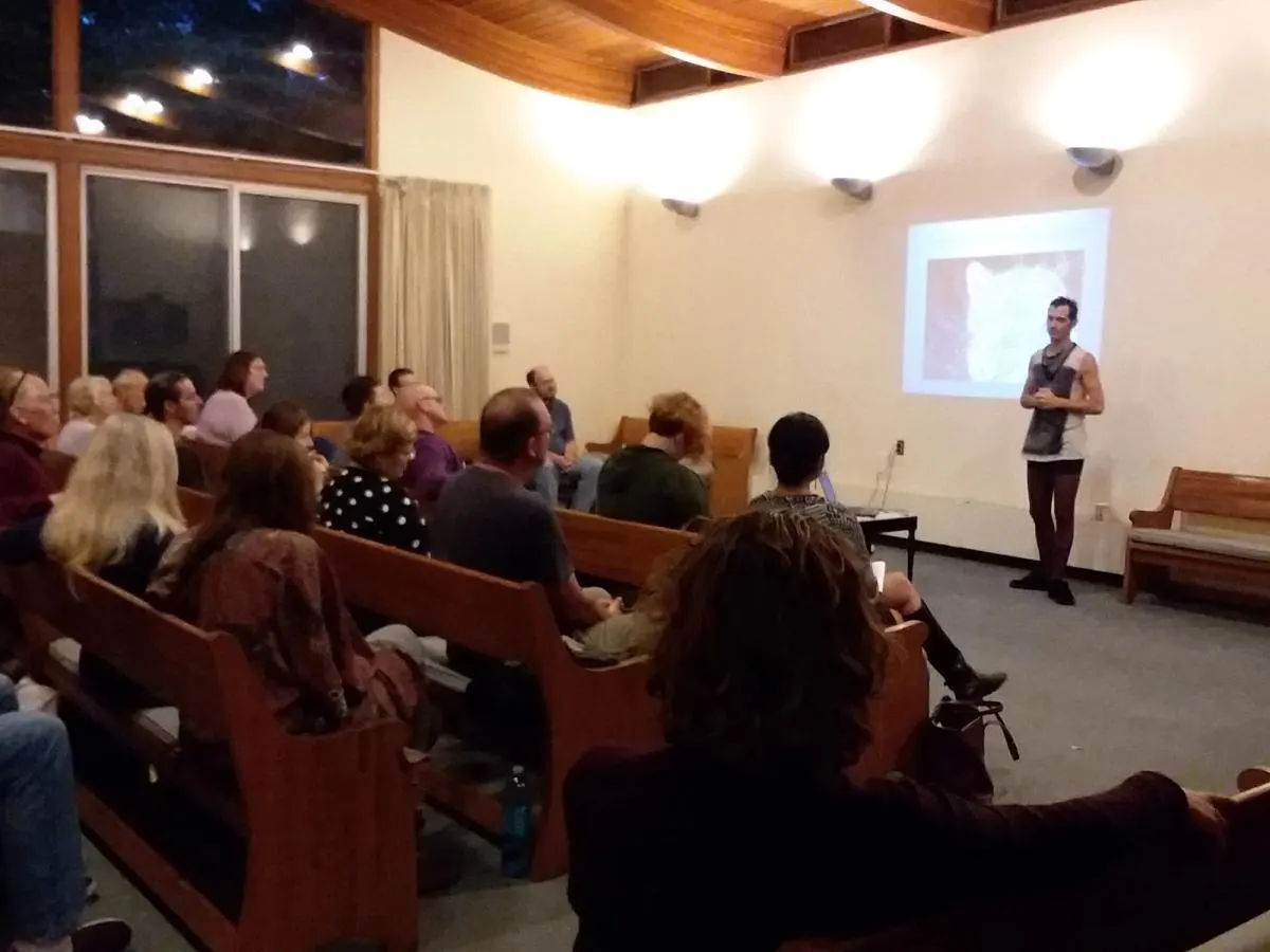 An audience in wooden pews listens to a speaker standing beside a projection screen.