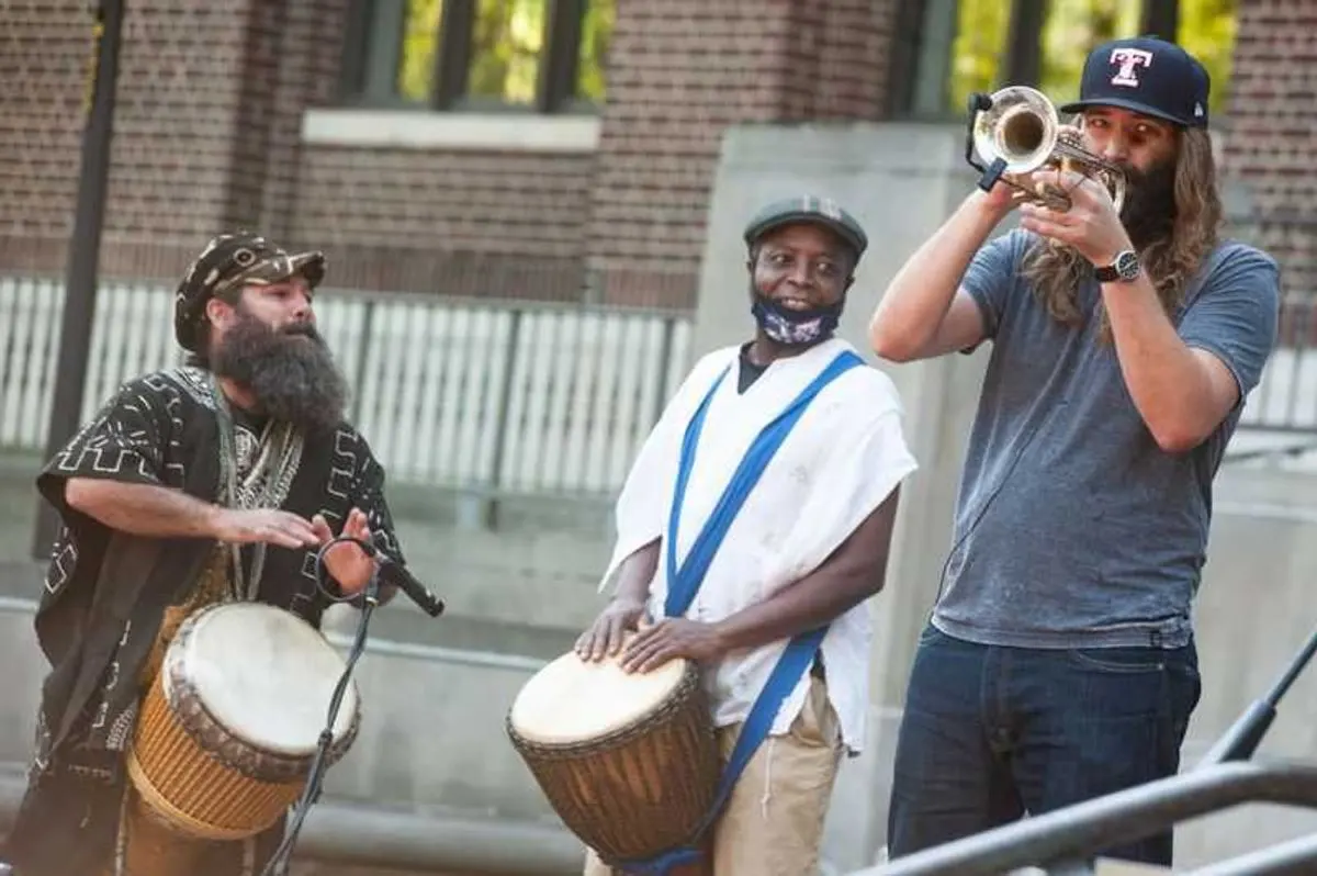 Three musicians play djembe drums and a trumpet outside a brick building.