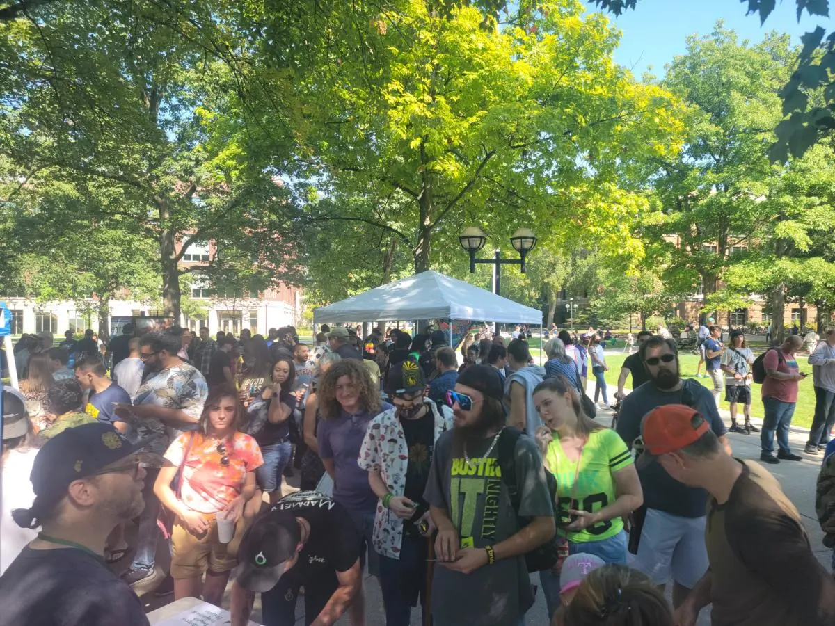 A summer crowd gathers under green trees on the Diag beside a pop-up tent.