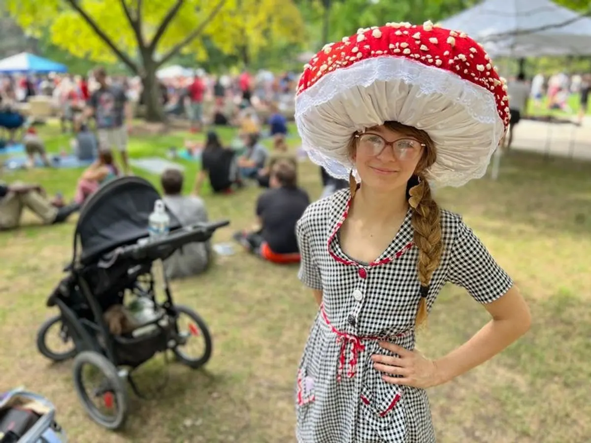 A festival goer in a giant red and white mushroom hat stands on the grass, with the crowd picnicking behind.