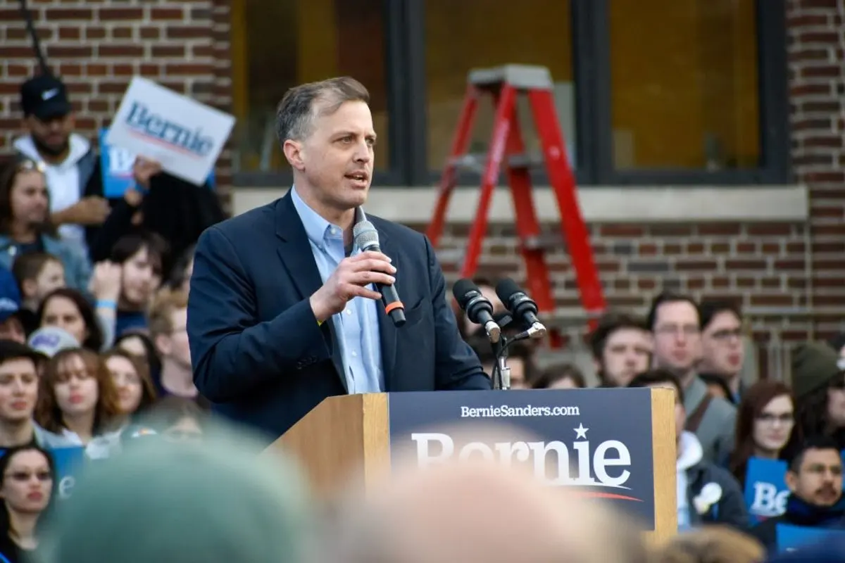 A speaker addresses a crowd from a podium crowded with microphones and campaign signs.
