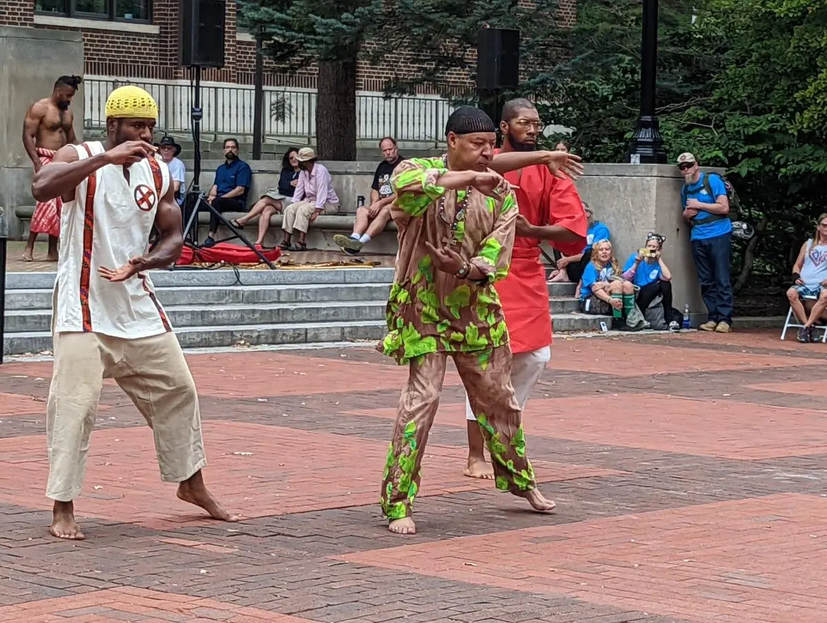 Dancers in bright patterned dress perform on the brick paving of the Diag while people watch from the steps.